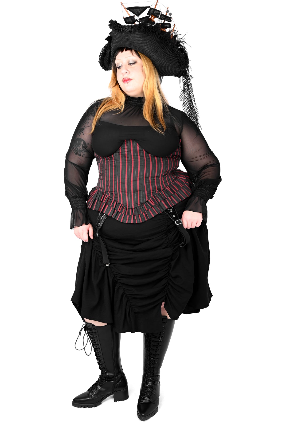 A woman modeling a gothic pirate costume against a white background. The outfit features a black sheer long-sleeve top, a red and black striped corset, a black ruffled skirt, black lace-up boots, and a black pirate hat with a skull and crossbones.