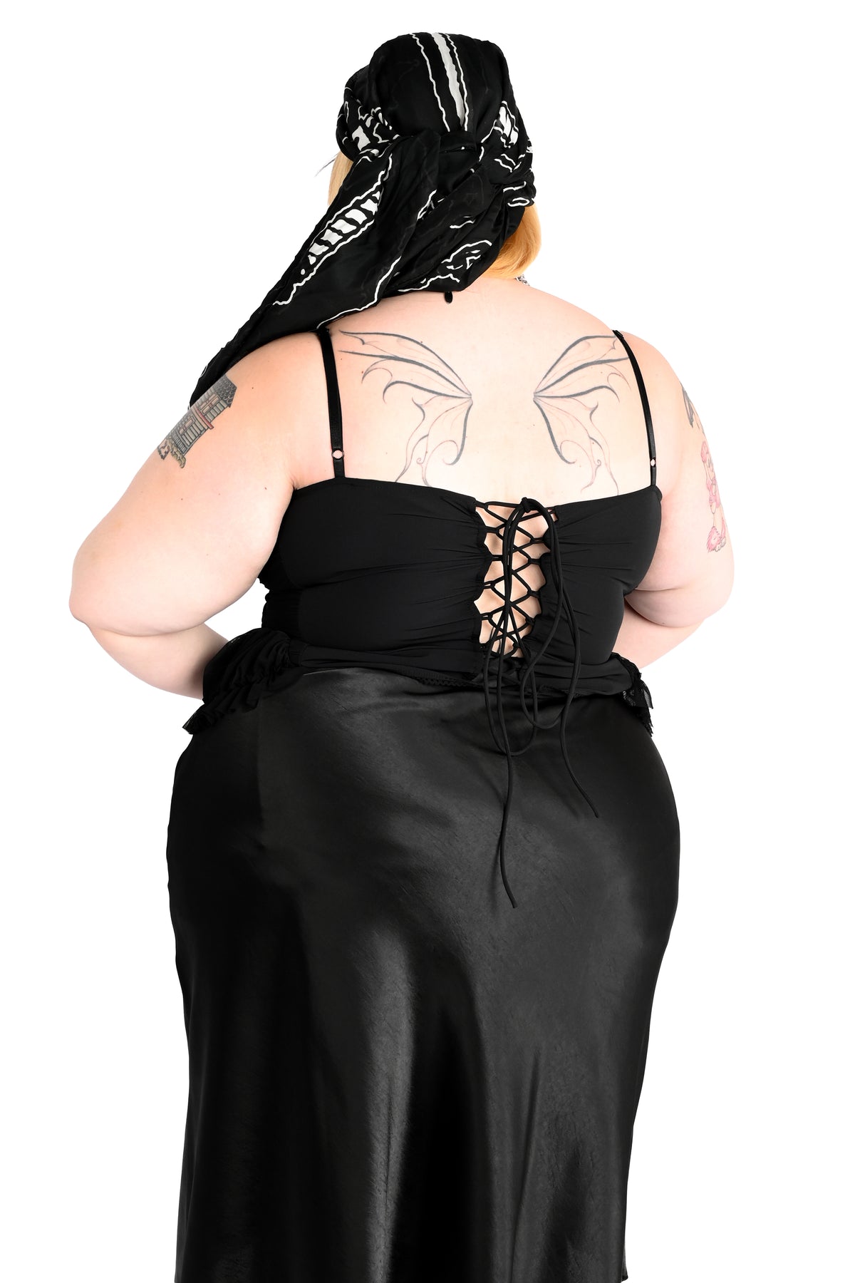 A person shown from the back wearing a black satin-like dress with thin straps and a lace-up back, along with a black patterned scarf. The background is plain white.