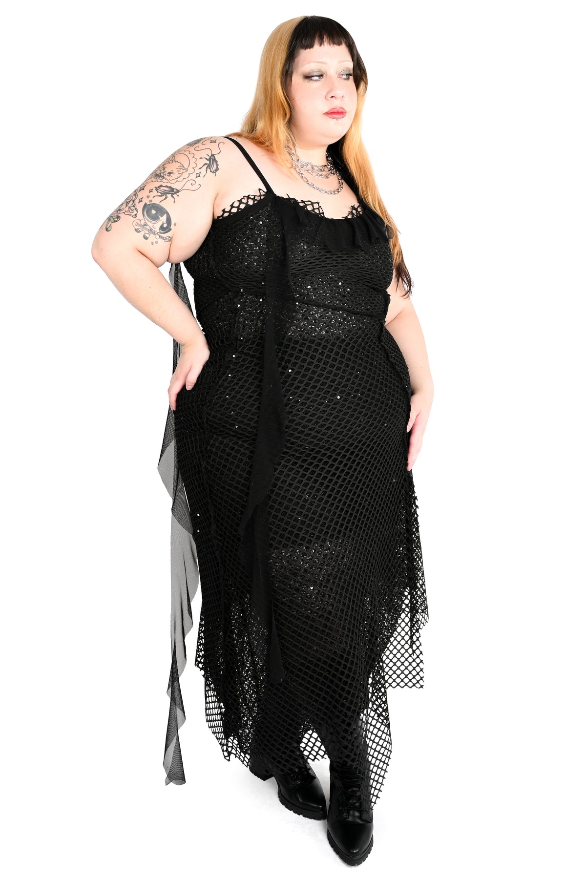 A plus-size model with blonde hair and tattoos wearing a black fishnet midi dress with spaghetti straps, a ruffled sweetheart neckline, and a sheer side drape, photographed against a solid white background.