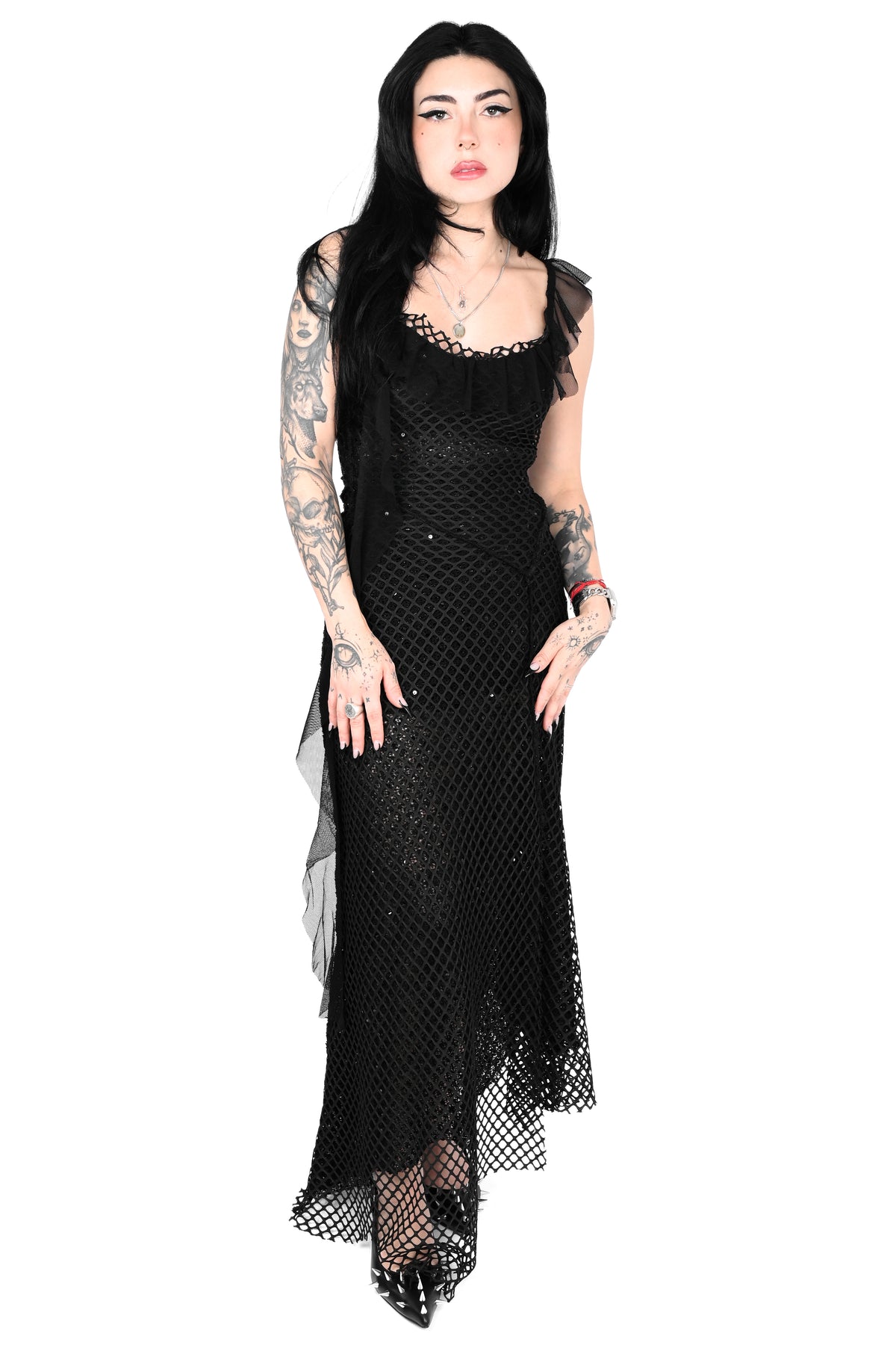 A model with long black hair and visible arm tattoos wears a long black fishnet dress with sheer mesh sleeves and a high-low hem, posed against a solid white background.