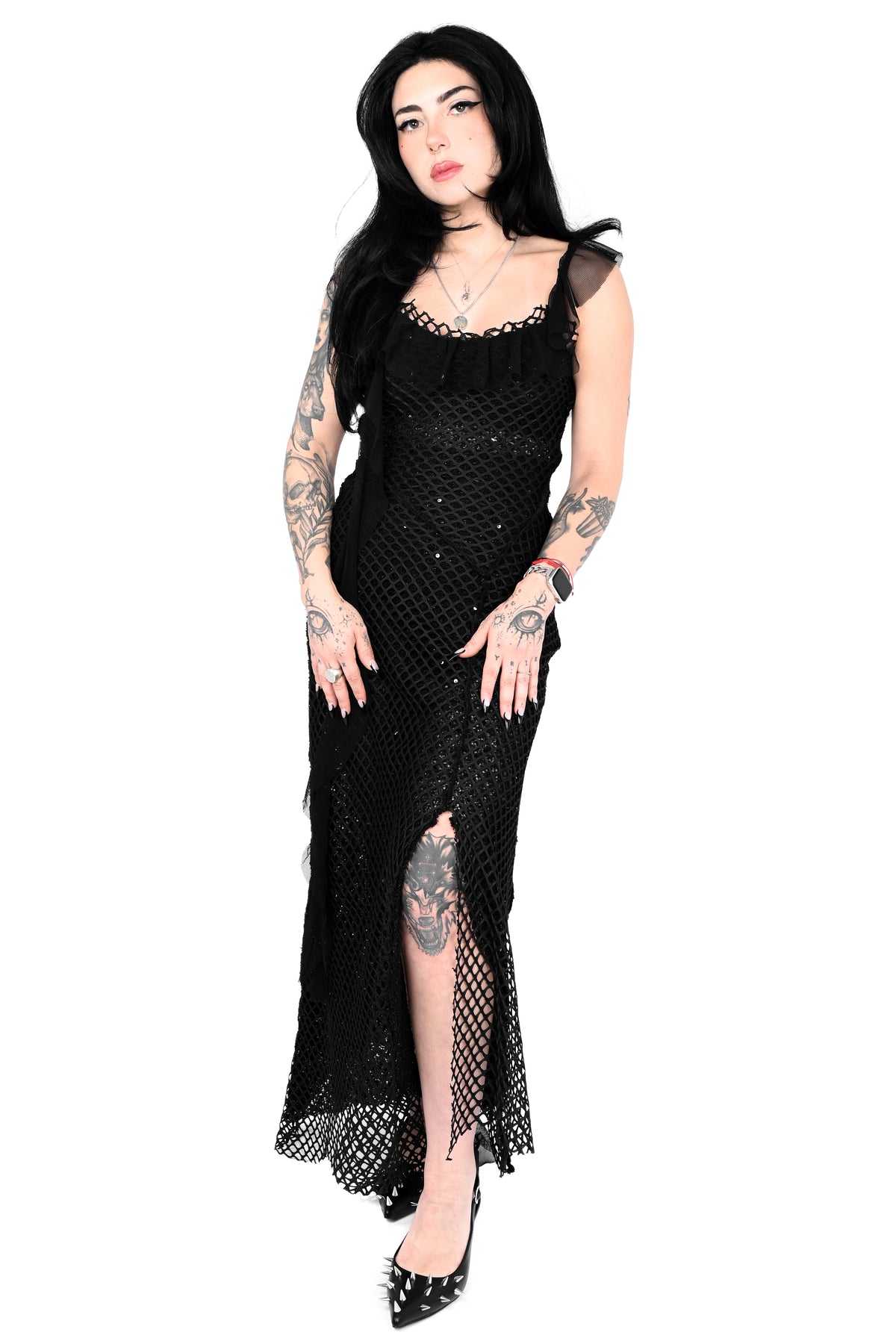 A model with tattoos wearing a black crochet knit maxi dress with sheer shoulder straps, a ruffled neckline, and a high slit, paired with black studded heels against a white background.