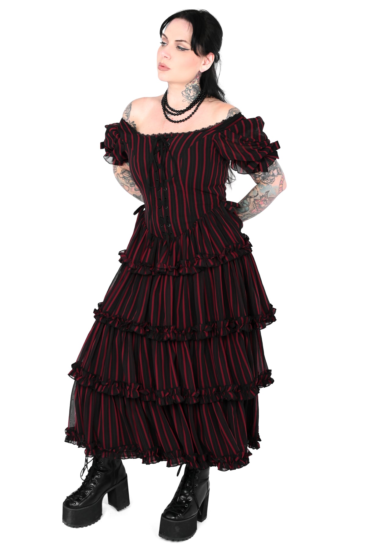 A model with dark hair and visible arm tattoos wears a black off-the-shoulder tiered dress with thin red vertical stripes, paired with black platform boots, against a solid white background.