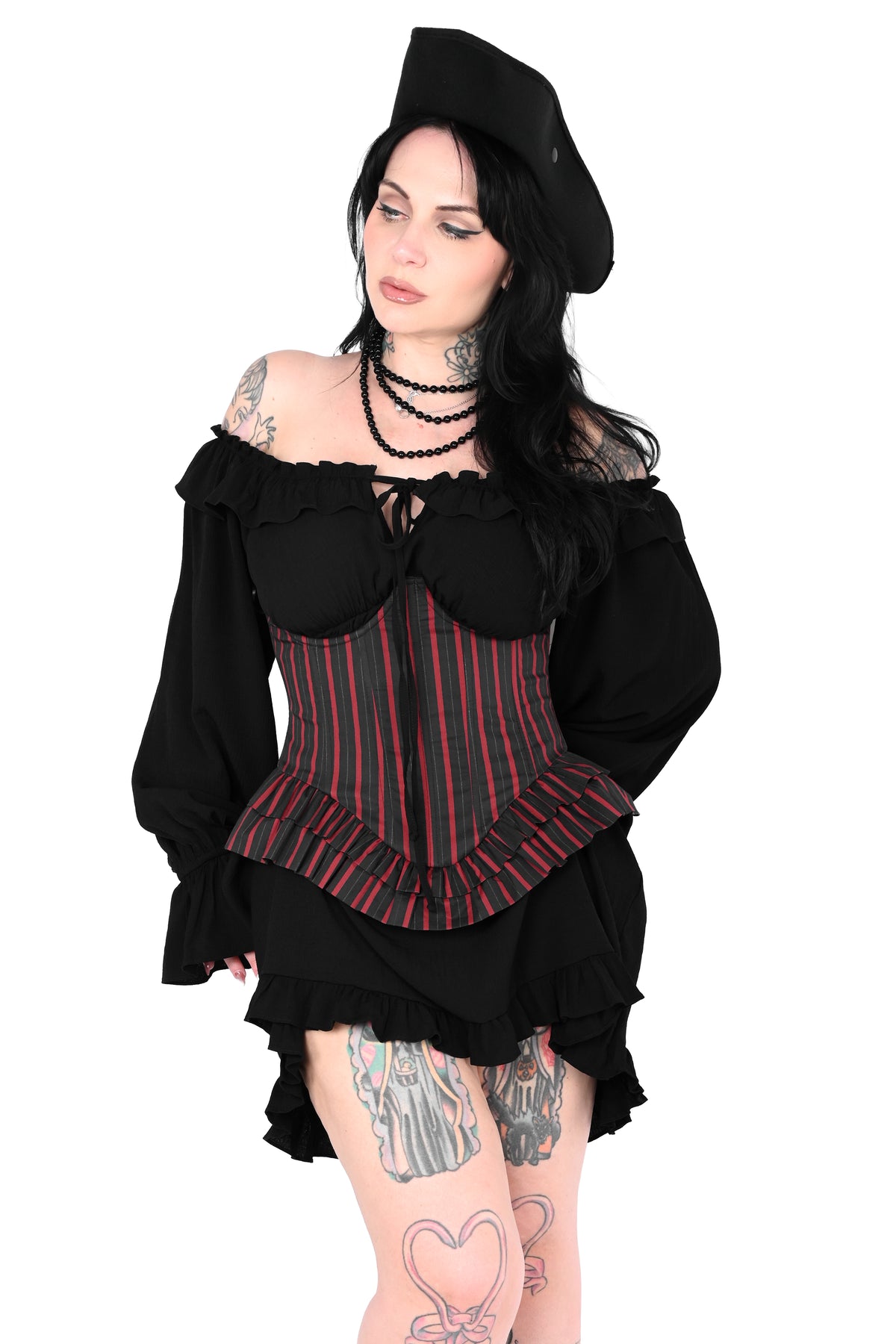 A woman modeling a black gothic-style costume top with red and black vertical stripes, a ruffled off-the-shoulder neckline, and a matching black hat against a plain white background.