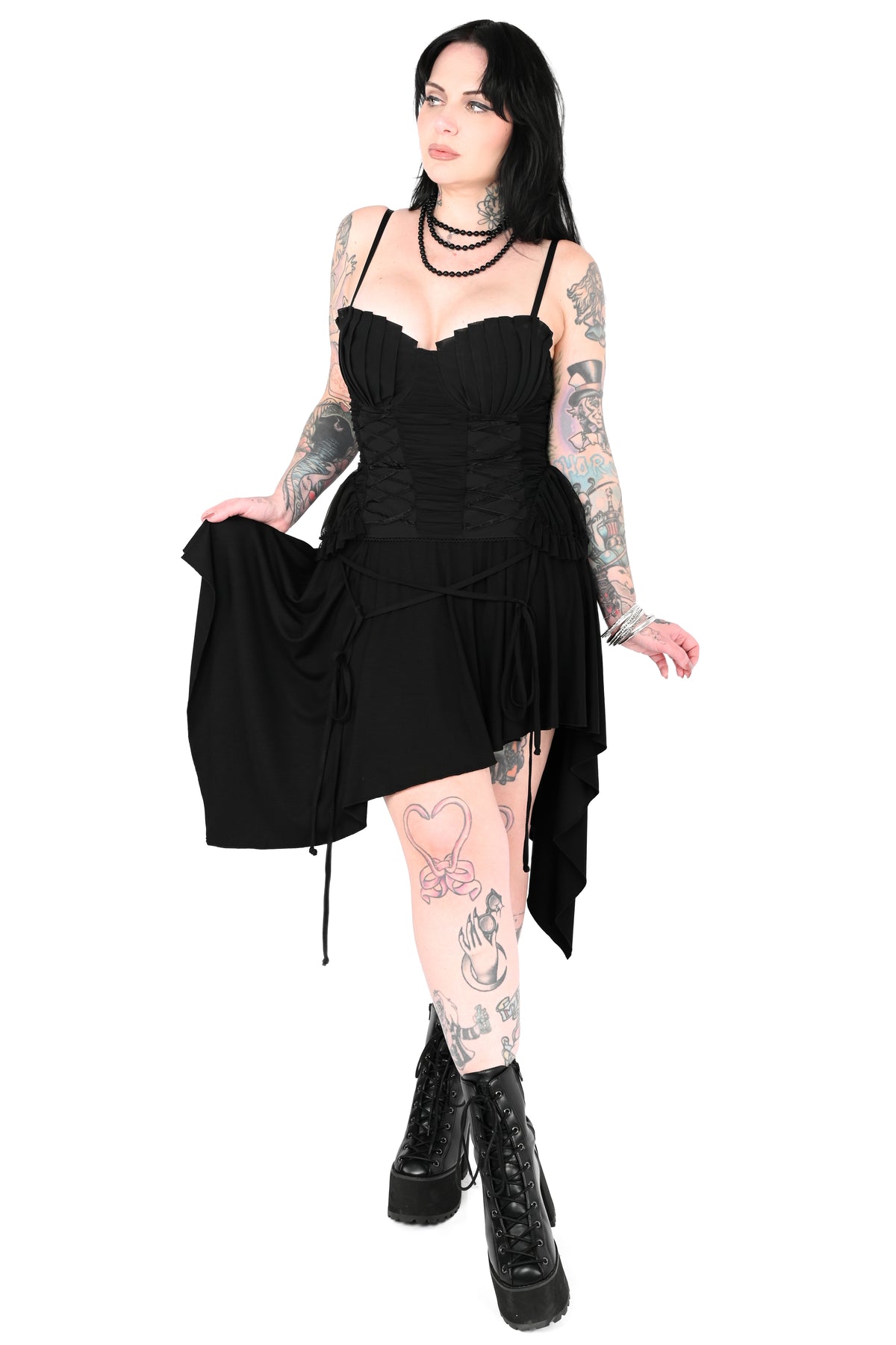 A woman with tattoos models a black asymmetrical high-low dress with a sweetheart neckline, corset-style bodice, and draped skirt, paired with black platform boots against a plain white background.