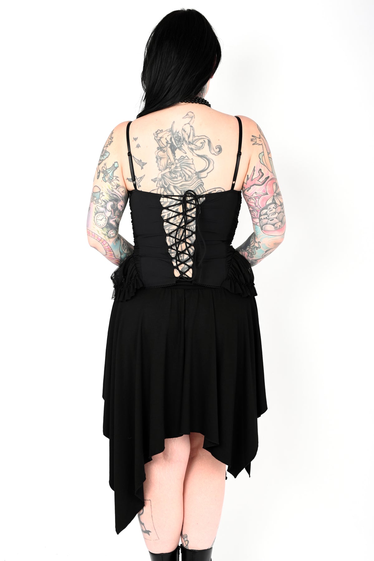 A person shown from the back wearing a black asymmetrical high-low dress with thin straps, a lace-up back, and ruffled trim, against a plain white background.