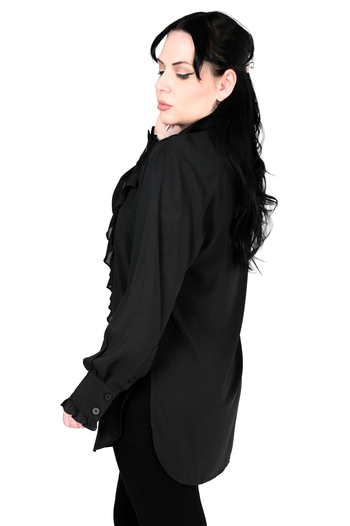 A woman with long black hair is shown in profile wearing a black long-sleeve blouse with ruffle detailing on the front and cuffs, paired with black pants, against a solid white background.