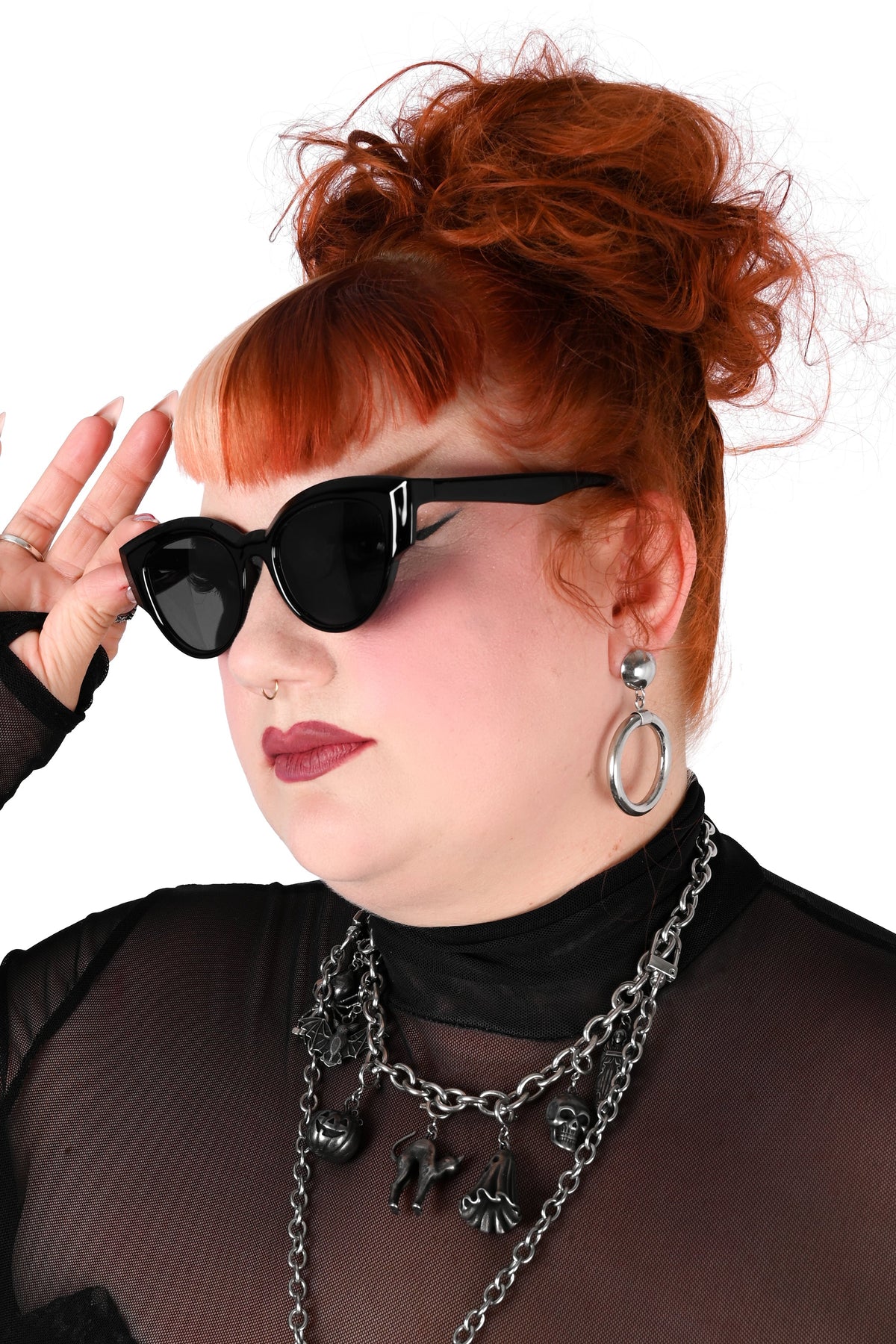 A woman with red hair in a messy bun wearing black cat-eye sunglasses, a sheer black long-sleeve top, layered silver chain necklaces, and silver hoop earrings against a white background.
