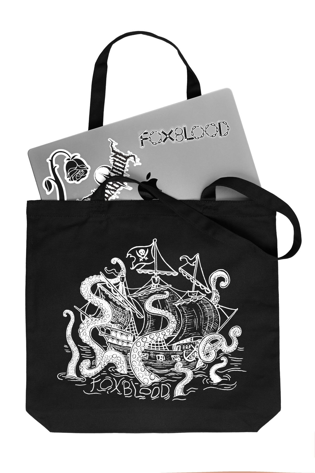 A silver Macbook peeking out of a black canvas tote bag with black handles, featuring a white line art graphic of a pirate ship being swallowed by a giant sea monster, with the text 'Foxblood' written /below in the ocean water lines. Shot on a white background.