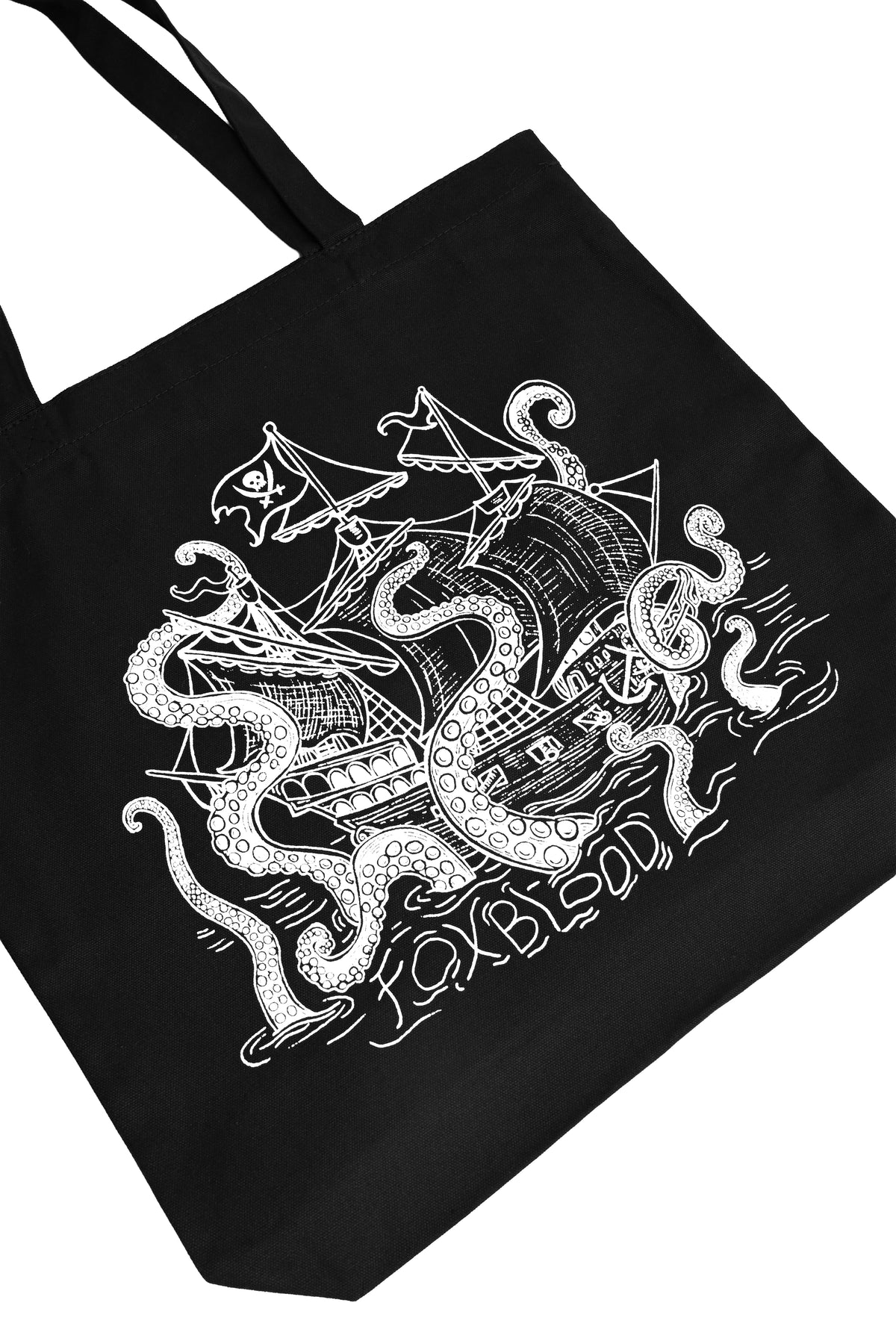 A close up angled flatlay photo of a black canvas tote bag with black handles, featuring a white line art graphic of a pirate ship being swallowed by a giant sea monster, with the text 'Foxblood' written /below in the ocean water lines