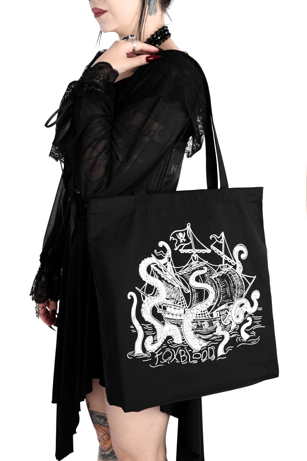 A model holding a black canvas tote bag on her shoulder. The bag has a white sea creature attacking a pirate ship with the letters "FOXBLOOD" spelled in the water below. Shot against a plain white background.
