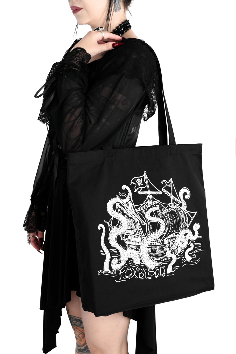 A model holding a black canvas tote bag on her shoulder. The bag has a white sea creature attacking a pirate ship with the letters "FOXBLOOD" spelled in the water below. Shot against a plain white background.