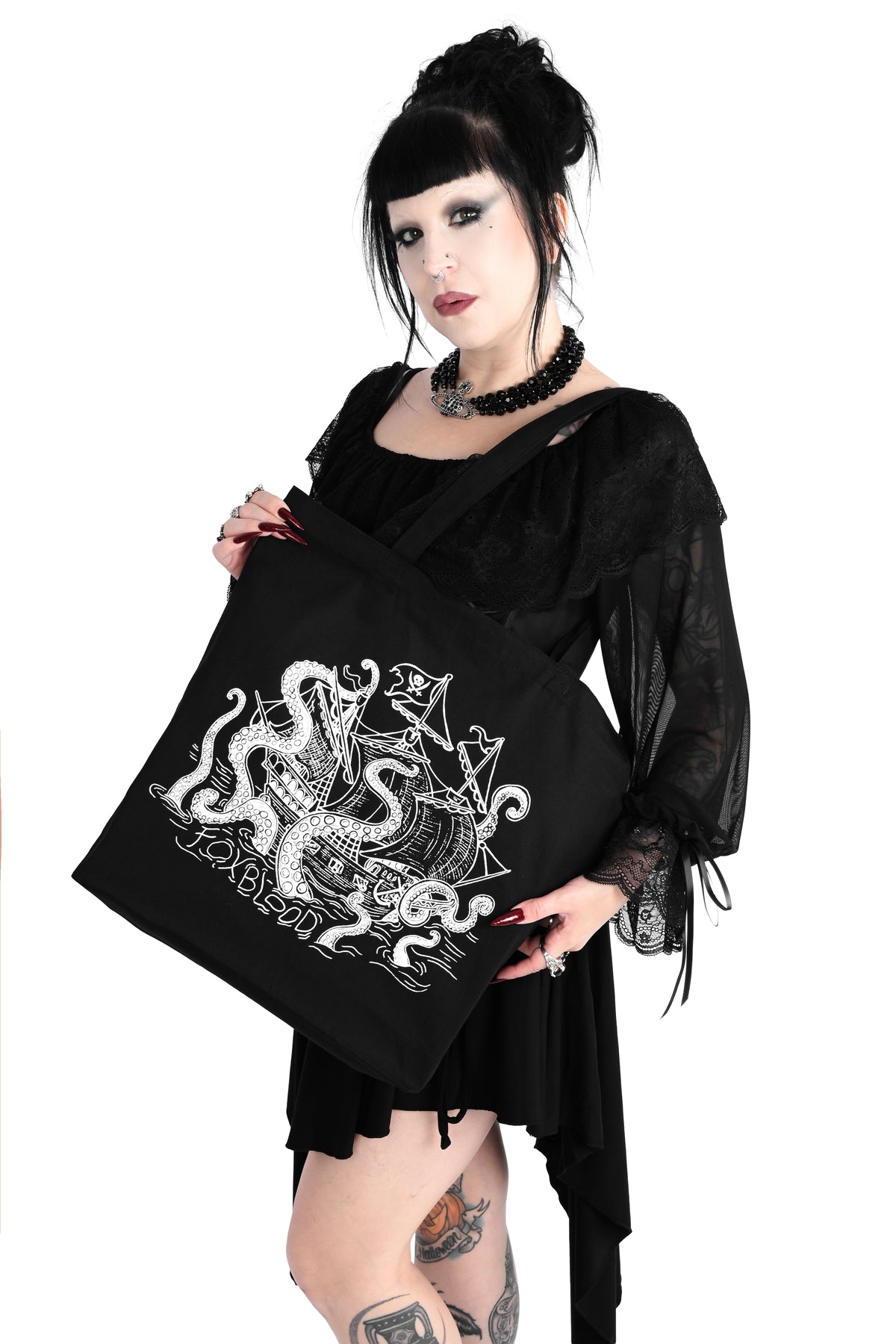 A slightly angled photo of a model holding a black canvas tote bag on her shoulder. The bag has a white sea creature attacking a pirate ship with the letters "FOXBLOOD" spelled in the water below. Shot against a plain white background.
