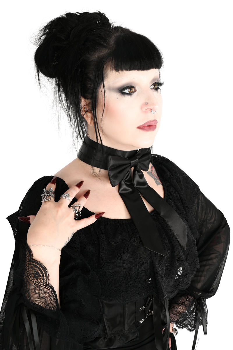 A 3/4 angle photo of a woman with dark hair in a bun wearing a black sheer long-sleeve blouse showing off a satin bow choker with long tails photographed against a plain white background.