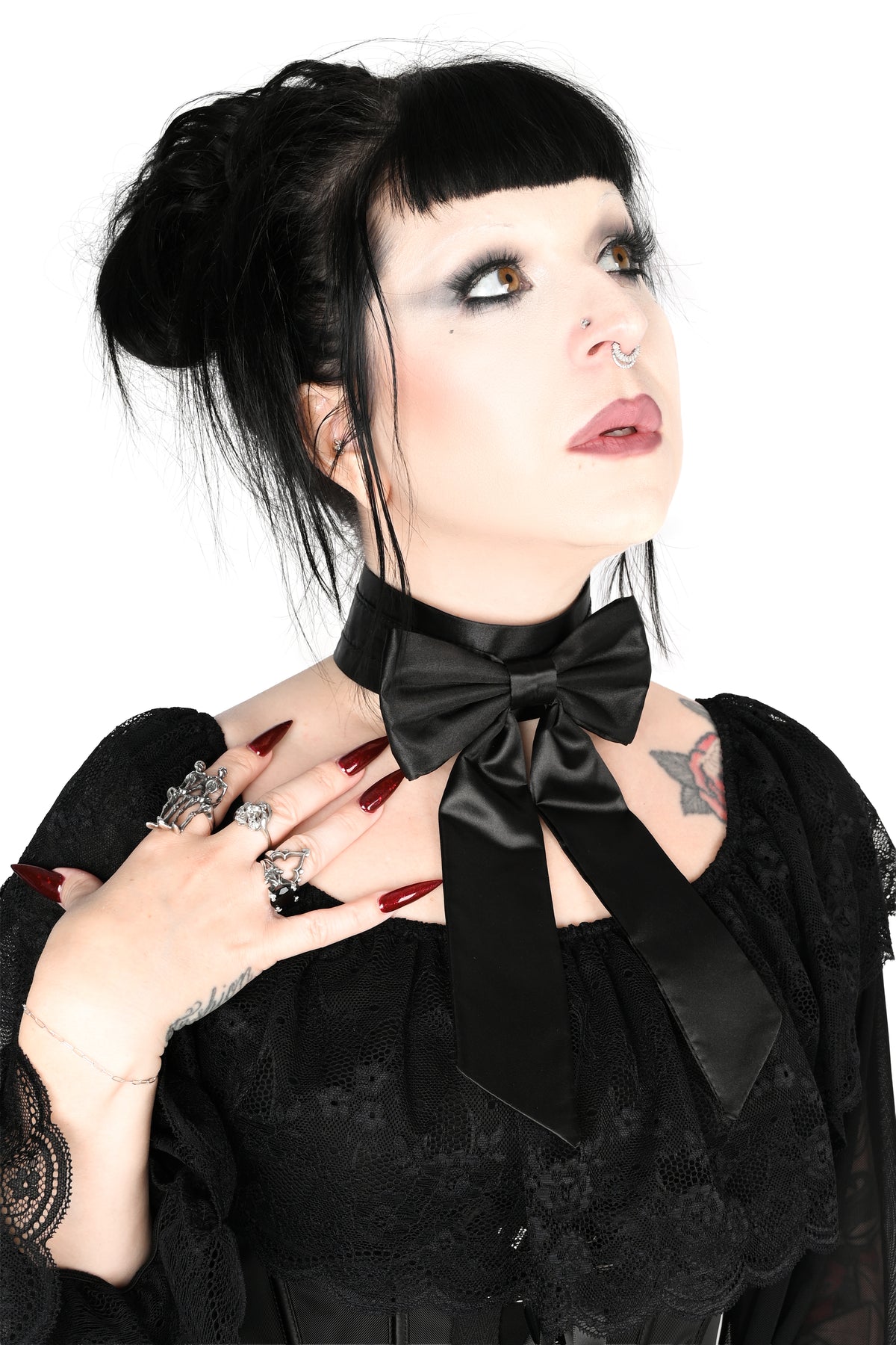 A  photo of a woman with dark hair in a bun looking up and to the side, wearing a black sheer long-sleeve blouse showing off a satin bow choker with long tails photographed against a plain white background.