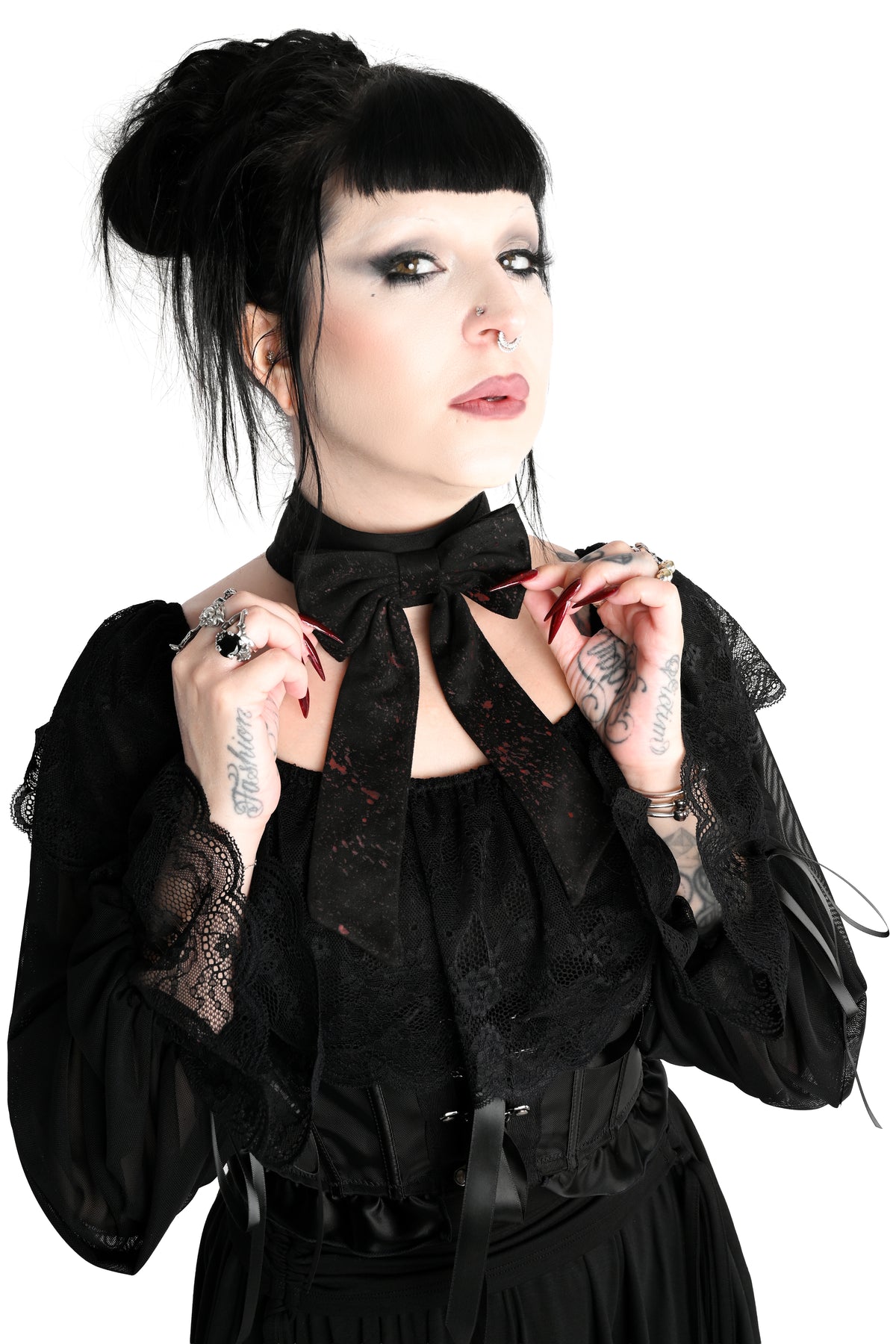 A woman with gothic styling, including a black topknot, dark eye makeup, and tattoos, wearing a black lace long-sleeve blouse showing off a large black bow choker with long tails and an all over splatter print, photographed against a solid white background.