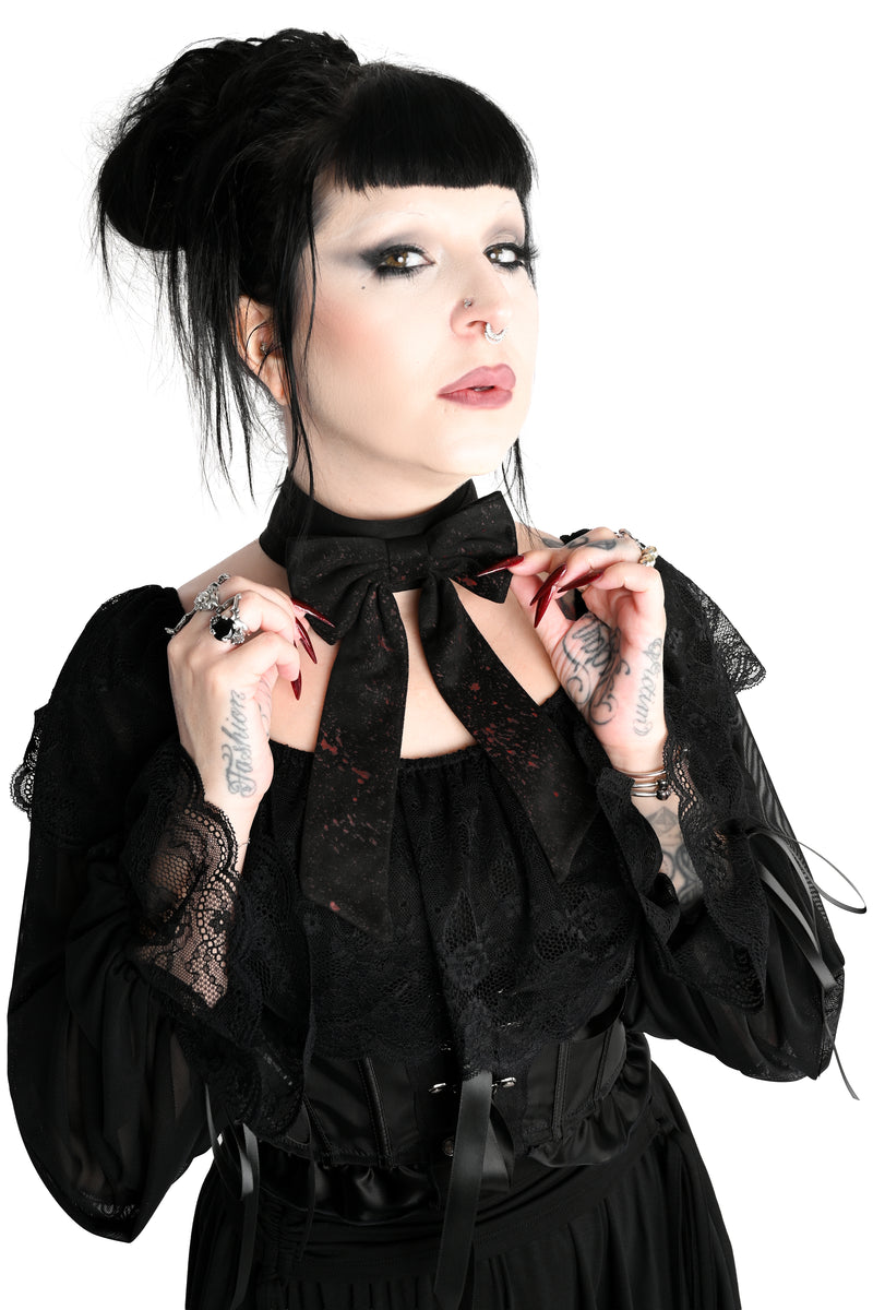 A woman with gothic styling, including a black topknot, dark eye makeup, and tattoos, wearing a black lace long-sleeve blouse showing off a large black bow choker with long tails and an all over splatter print, photographed against a solid white background.