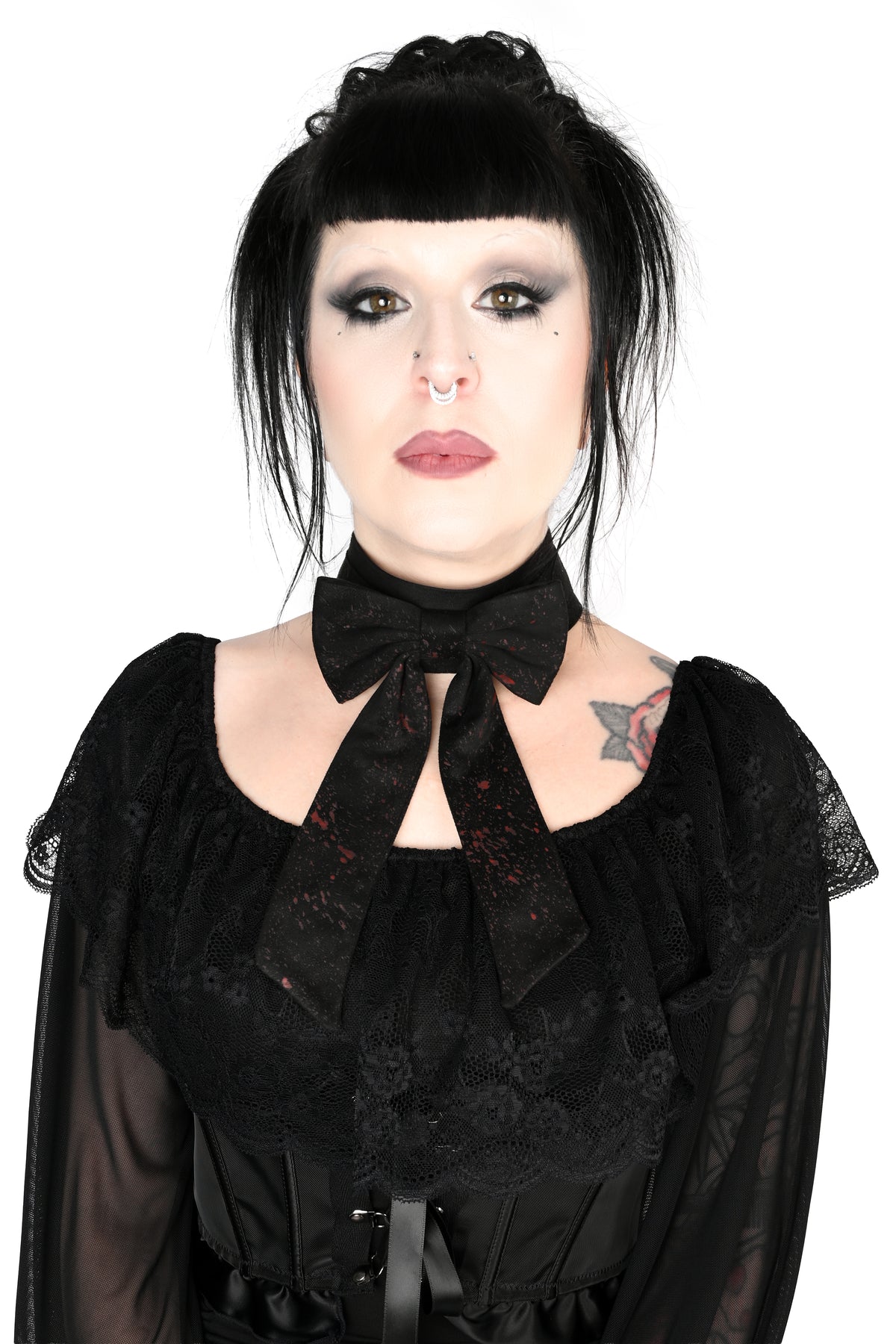 A fully front facing photo of a woman with gothic styling, including a black topknot, dark eye makeup, and tattoos, wearing a black lace long-sleeve blouse showing off a large black bow choker with long tails and an all over splatter print, photographed against a solid white background.