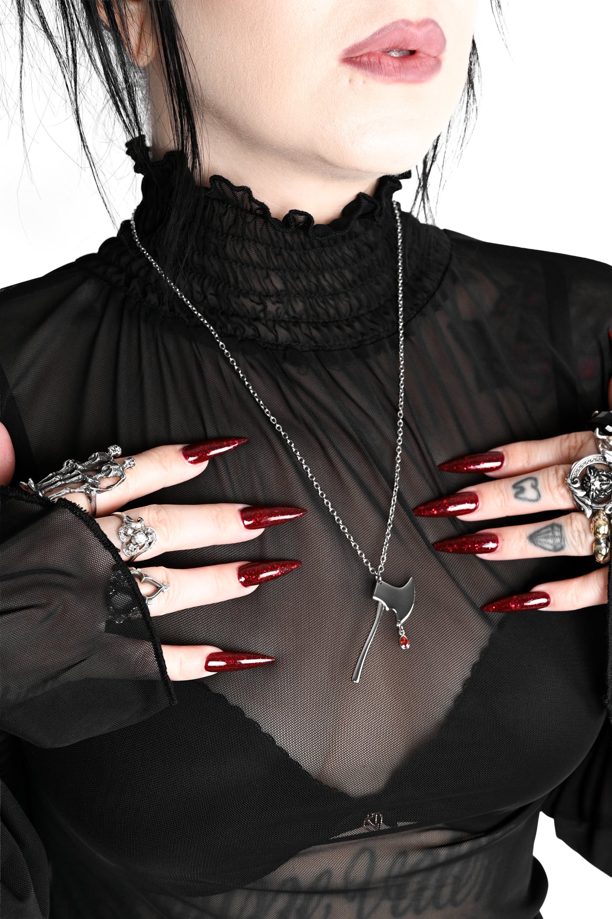 A close-up of a person wearing a black sheer long-sleeve top with a ruffled high neck, layered over a black bralette. The person is wearing a silver chain necklace with an axe pendant, multiple silver rings, and has long dark red manicured nails against a plain white background.