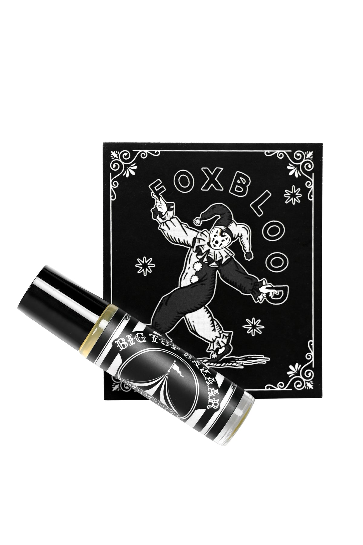 A black and white Foxblood glass rollerball perfume laid diagonally in front of its display card, which features a white jester illustration and the brand name 'FOXBLOOD'.
