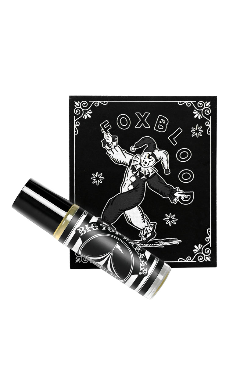 A black and white Foxblood glass rollerball perfume laid diagonally in front of its display card, which features a white jester illustration and the brand name 'FOXBLOOD'.