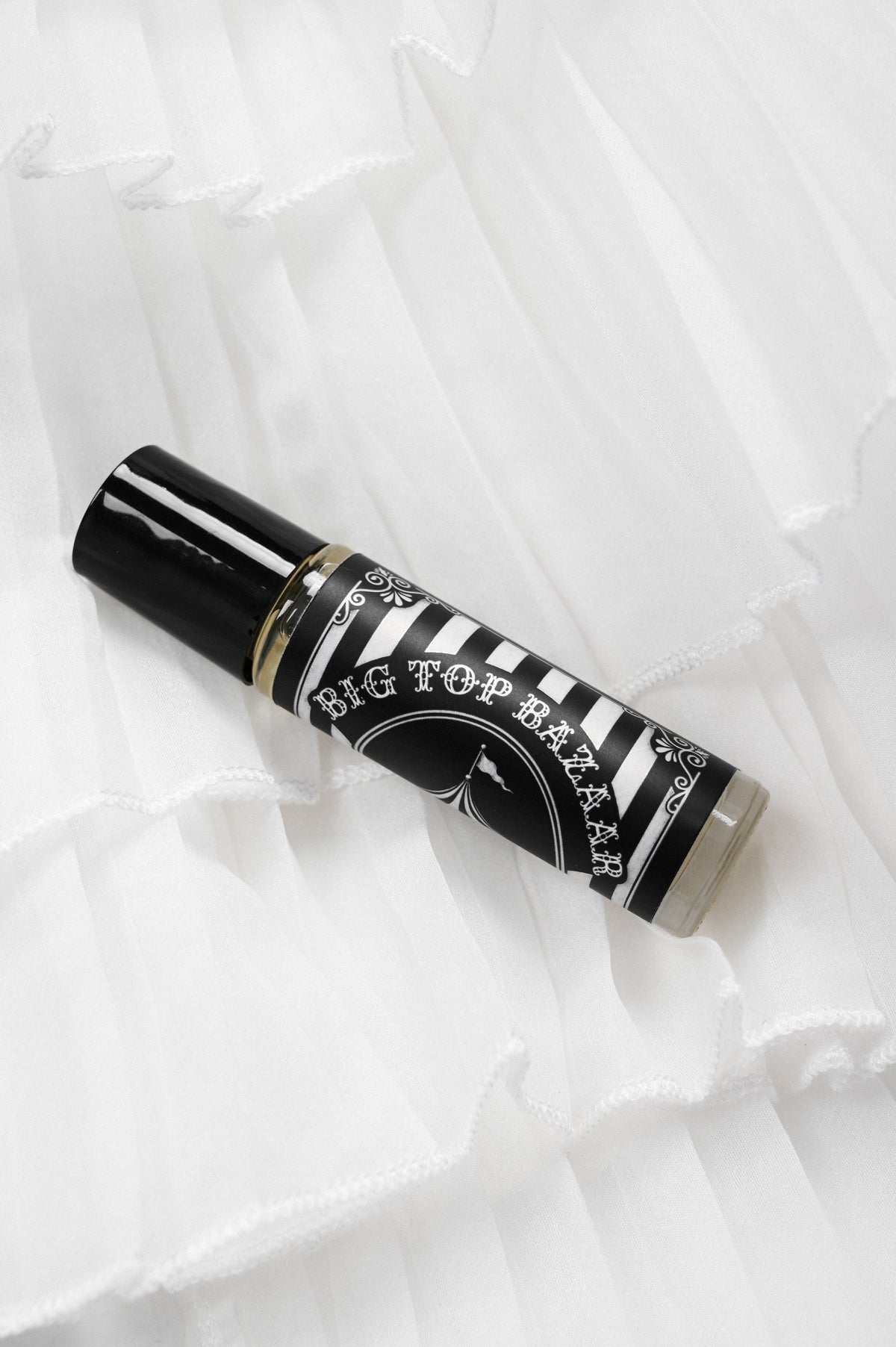 A glass roller ball perfume bottle with a black and white striped pattern and the brand name 'BIG TOP BAZAAR' printed on it, resting on a white ruffled fabric background.