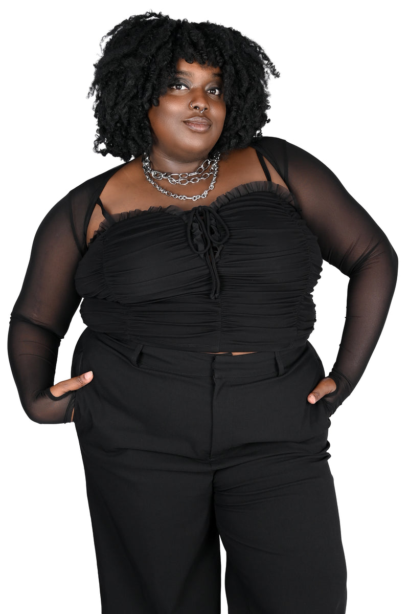 A plus-size model with curly black hair wearing a black top with a ruched sweetheart neckline and black pants, posed against a solid white background.