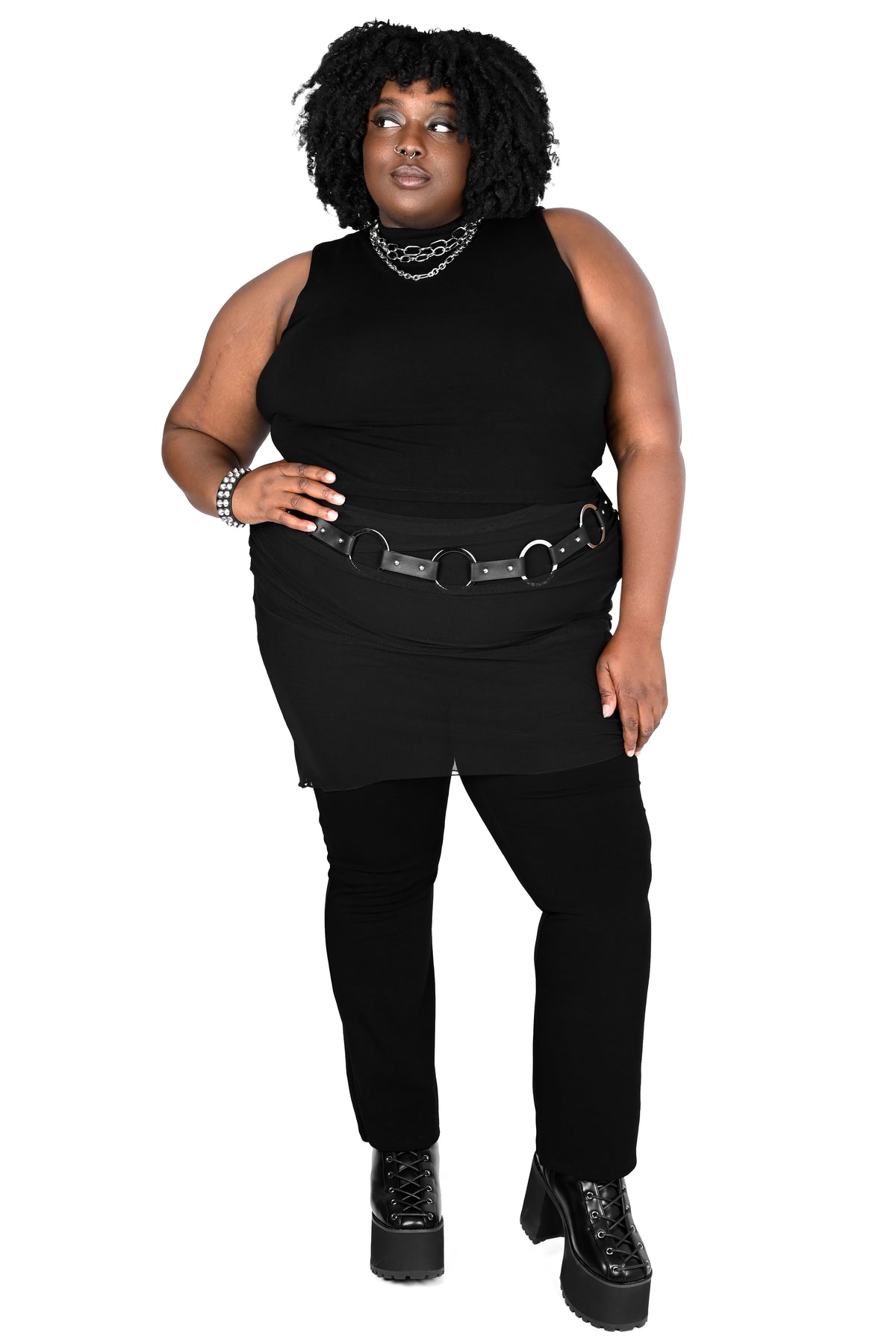 A plus-size model wearing a black sleeveless top, black mini skirt, black pants, and black platform boots, accessorized with a black chain belt and silver chain necklaces, against a plain white background.