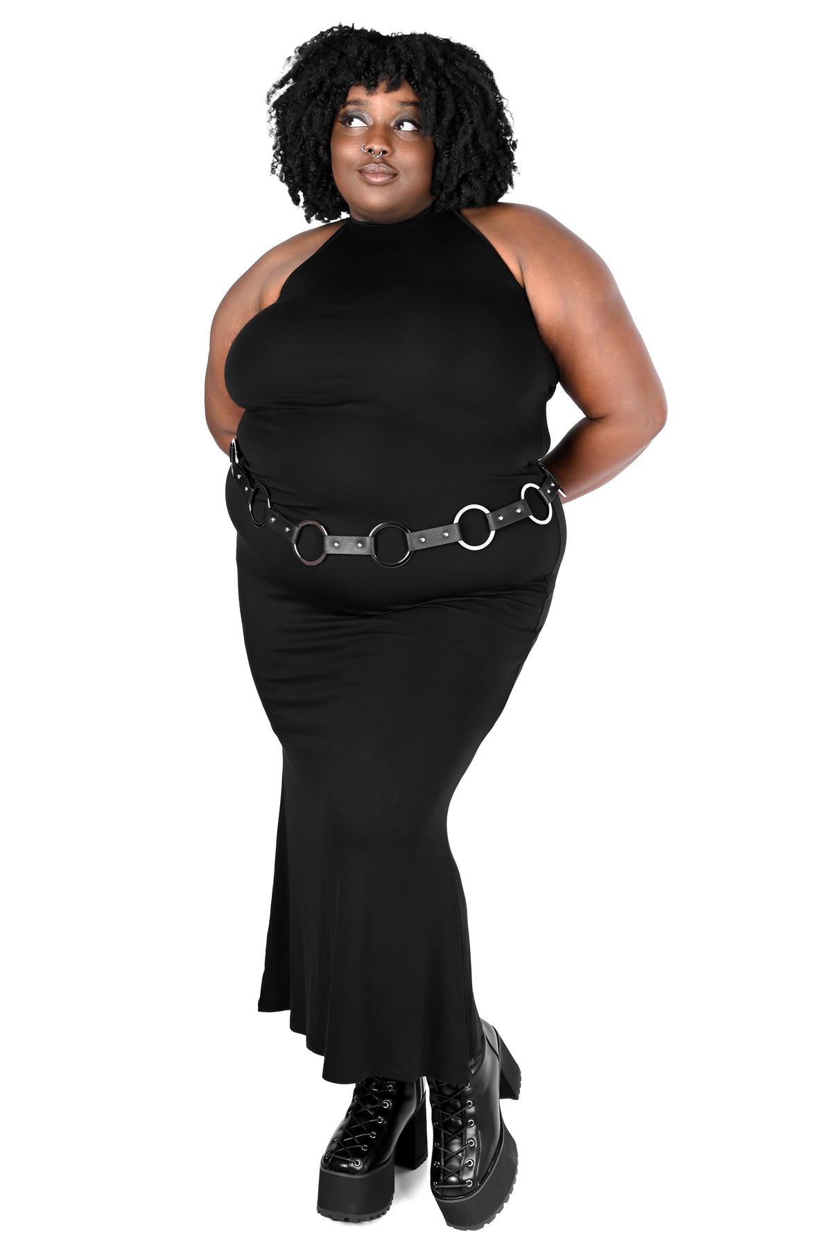 A plus-size model wearing a black halter-neck maxi dress with a black and silver o-ring belt, paired with black platform boots, against a solid white background.