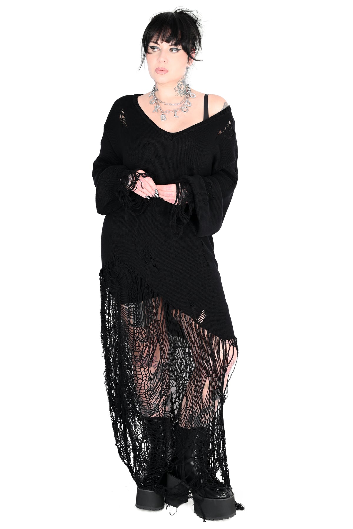 A model wearing a black distressed goth-style outfit featuring a fringed long-sleeve top, sheer lace hem, and platform boots against a plain white background.