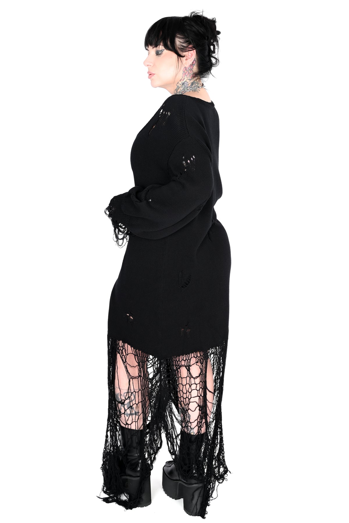A model shown from the side profile wearing a black long-sleeve distressed knit dress with a high-low hem, paired with black lace-up boots and sheer black fishnet tights.