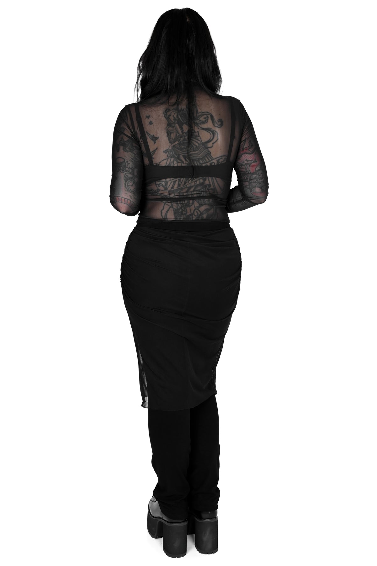 A woman viewed from the back wearing a black sheer long-sleeve mesh top, paired with a black wrap skirt and black platform boots, against a plain white background.