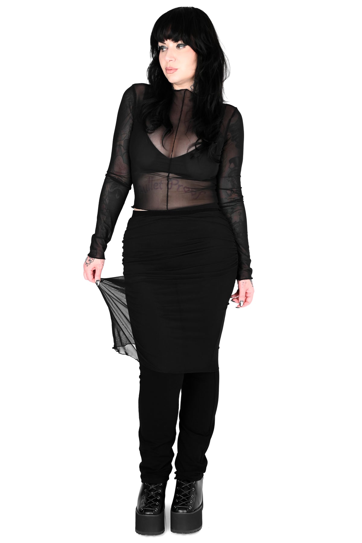 A woman modeling a black sheer long-sleeve crop top with a high neck, paired with a black ruched midi skirt, black pants, and black platform boots against a plain white background.