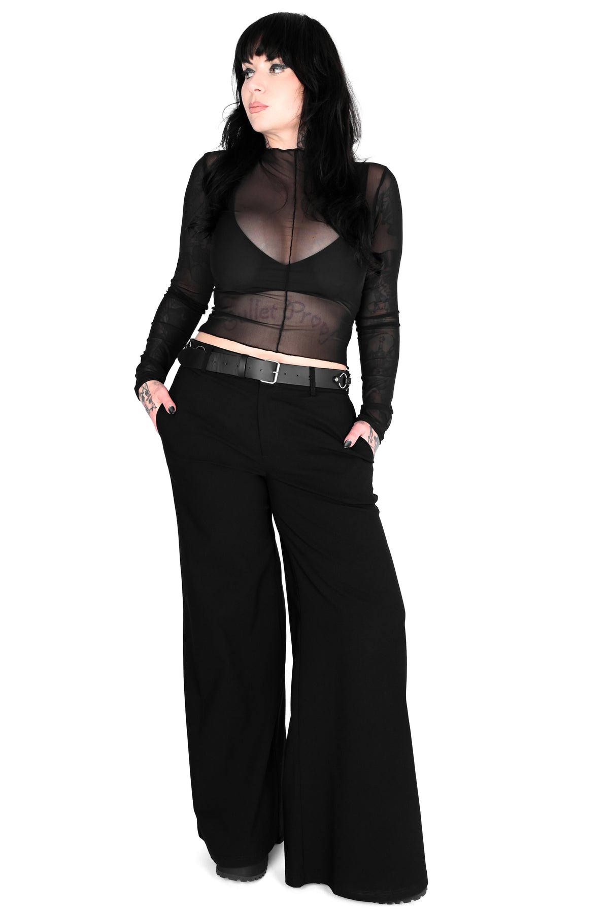A model with long black hair is wearing a black sheer long-sleeve crop top, a black bralette, a black belt, and wide-leg black pants, standing against a solid white background.
