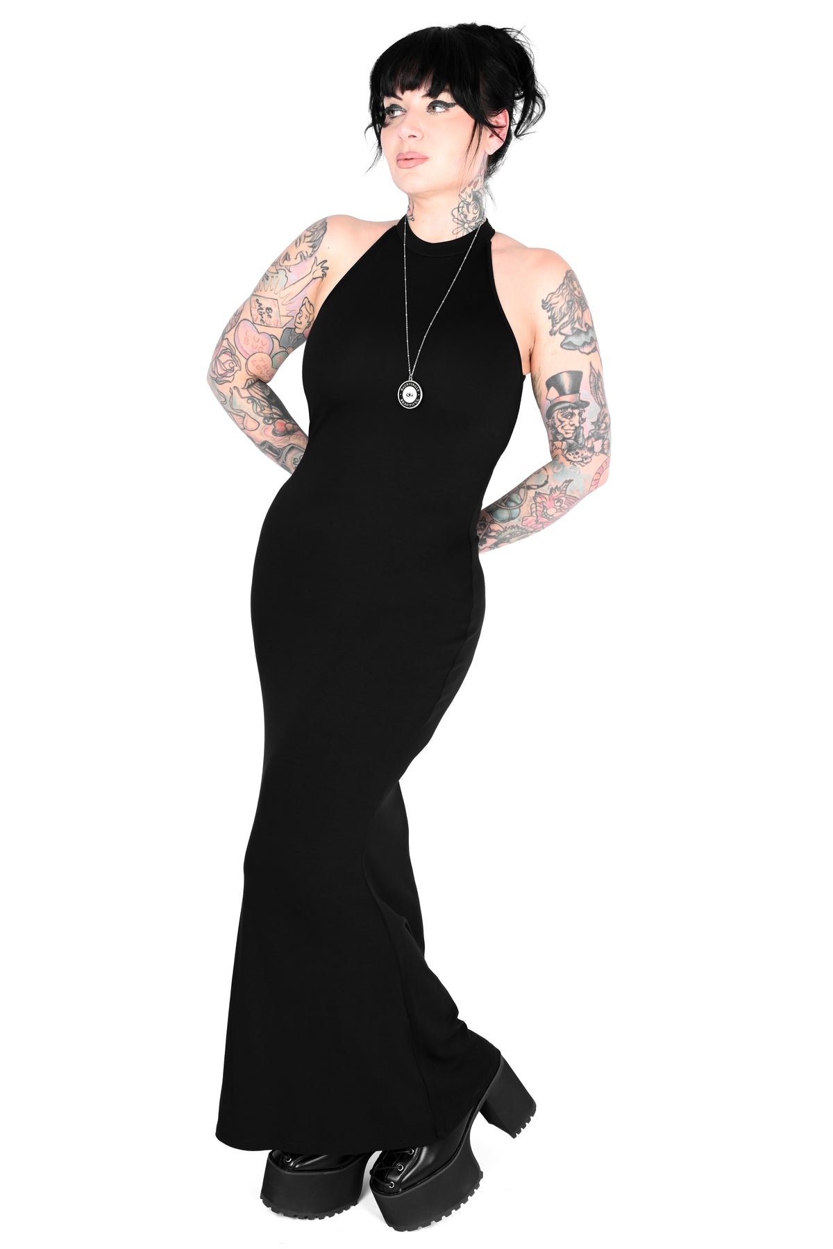 A model with black hair and visible tattoos wears a sleeveless, high-neck, floor-length black dress with a flared hem, paired with chunky black platform shoes and a silver pendant necklace, set against a plain white background.