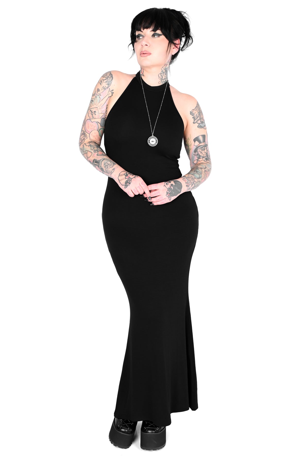 A woman with black hair and visible tattoos wears a fitted black halter-neck maxi dress with a flared hem in size medium, paired with black platform shoes and a silver pendant necklace, against a plain white background.