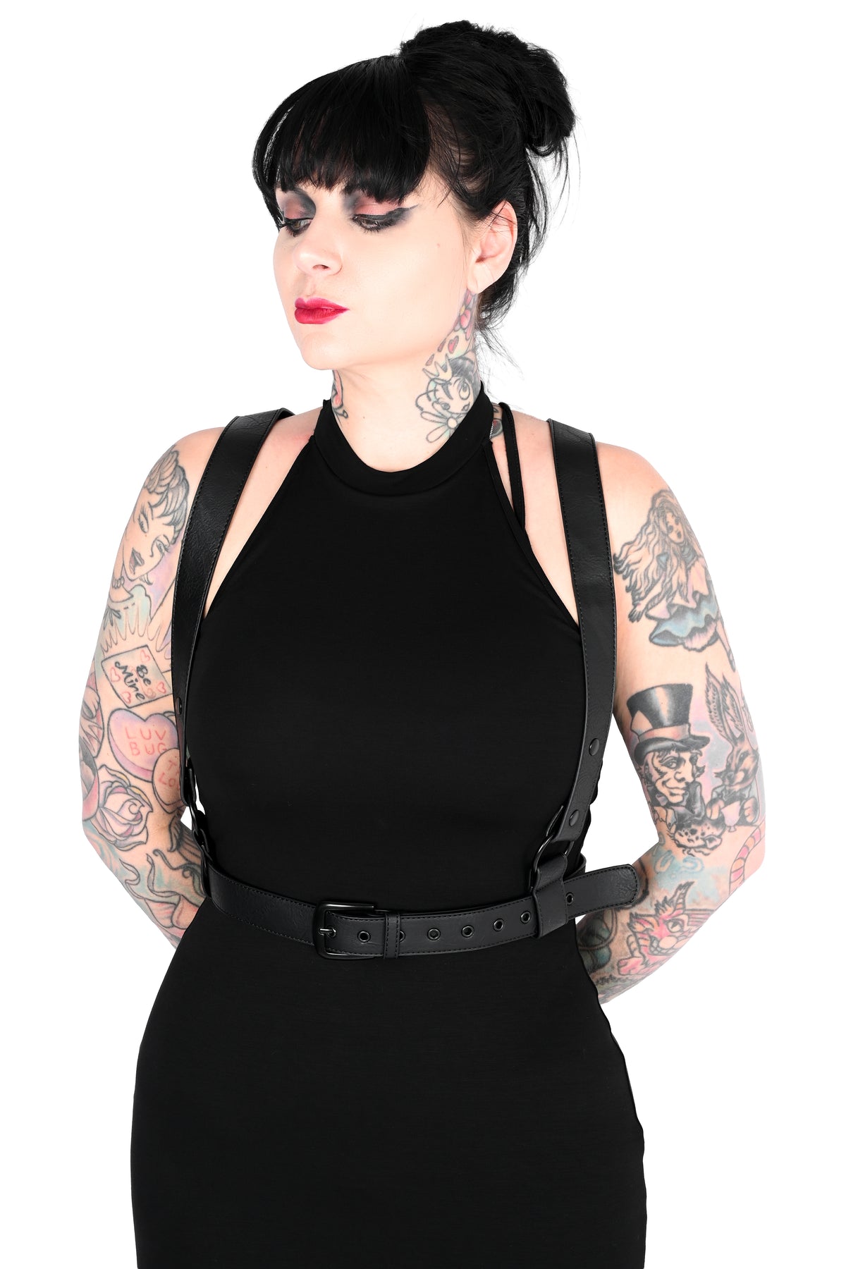 A woman with black hair in a bun and visible tattoos wears a black sleeveless dress with a harness against a plain white background.