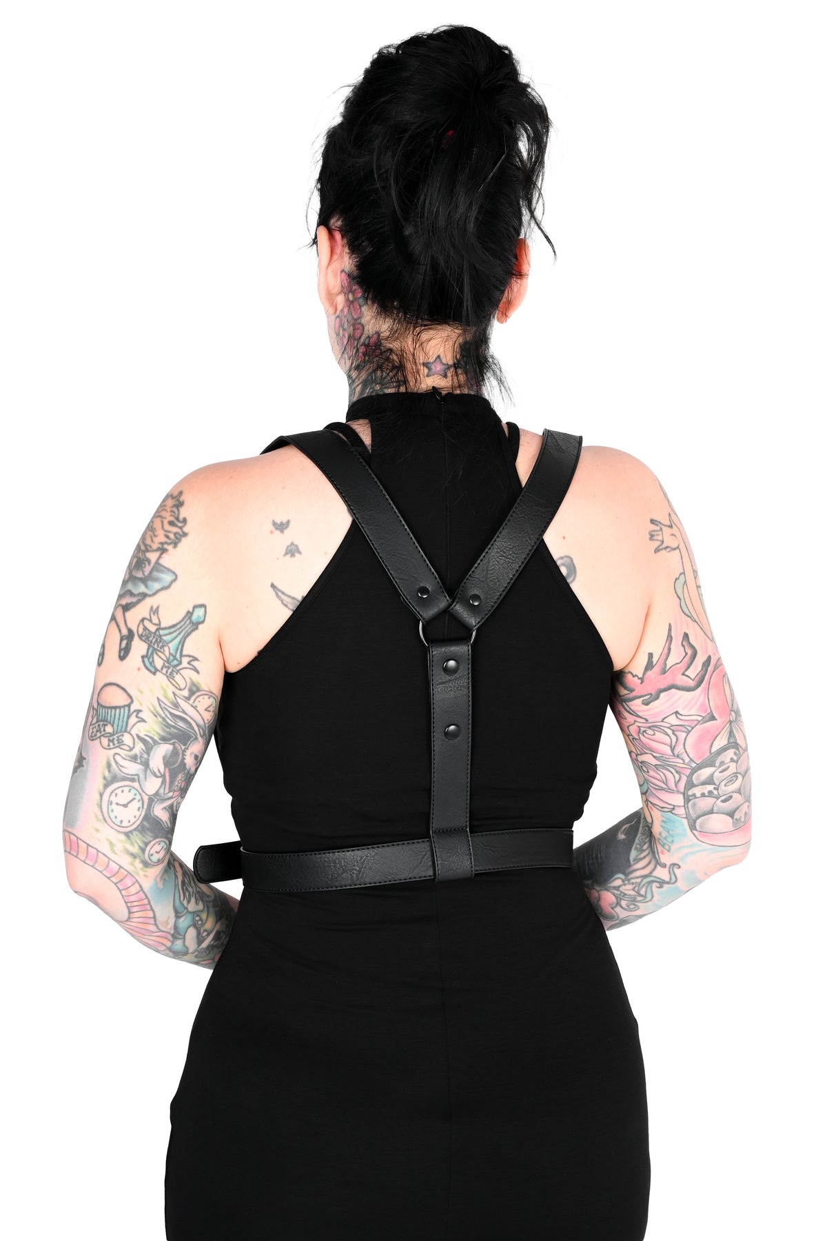 Back view of a tattooed woman with her hair in a bun, wearing a black dress and a black faux-leather harness-style strap accessory across her upper back and waist, against a plain white background.