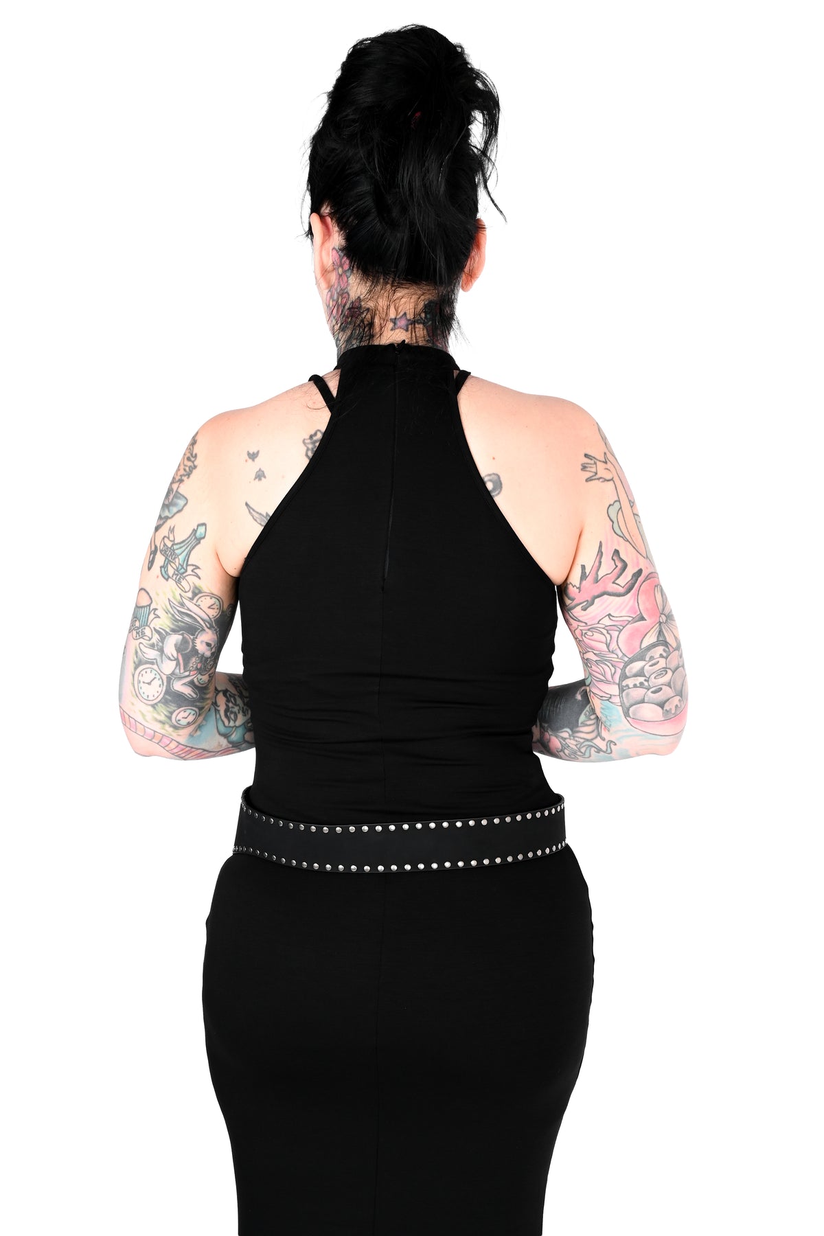 Back view of a person with dark hair in a bun, wearing a black halter-neck dress with a studded belt, against a plain white background.