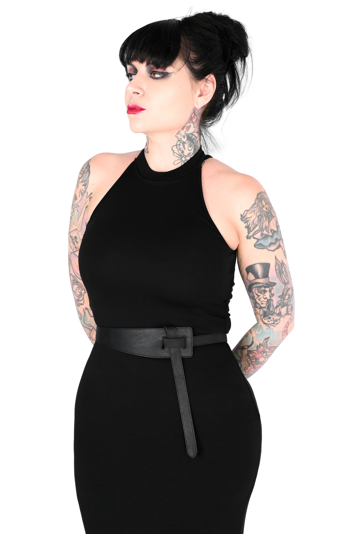 A woman with tattoos and black hair in a bun wearing a black sleeveless bodycon dress with a black wrap belt, photographed against a plain white background.