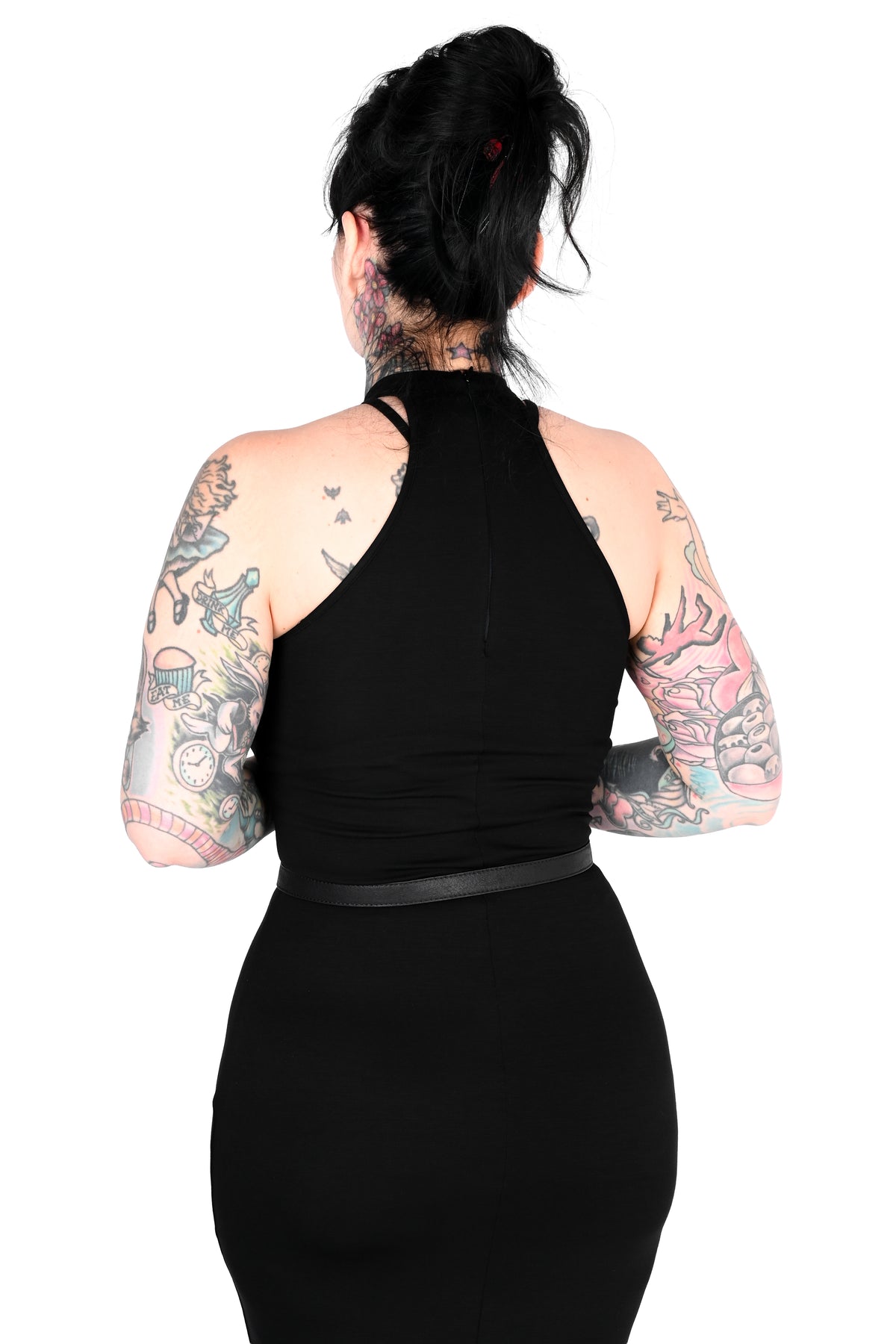 A woman with black hair in a bun and colorful tattoos on her arms, shown from the back wearing a black halter-neck dress with a thin black belt at the waist, against a solid white background.