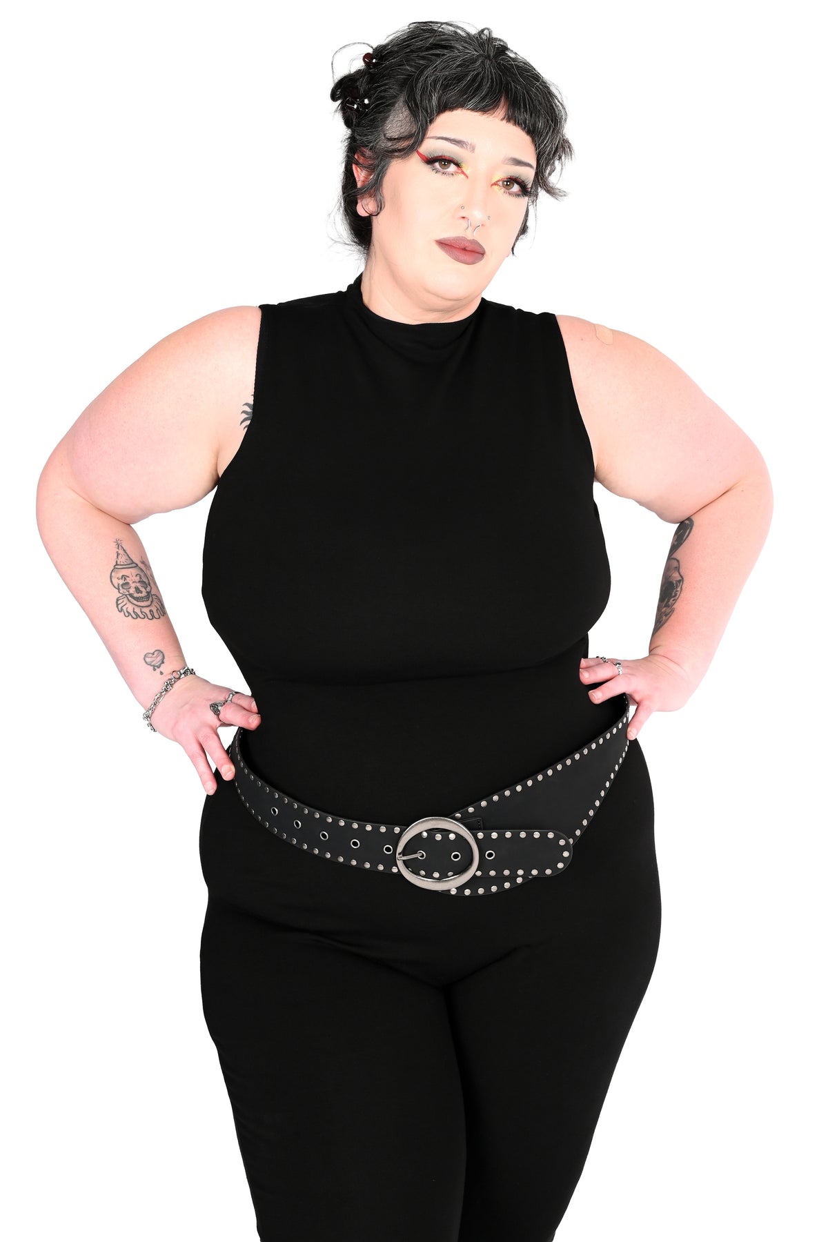 A plus-size model with short dark hair poses in a black sleeveless mock-neck jumpsuit wearing a black studded belt with a silver buckle, against a plain white background. Her arms are up on her hips and she is looking forward