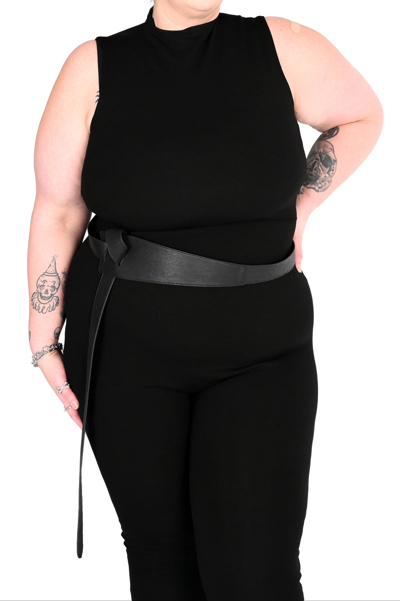 A plus-size model with short dark hair and tattoos wearing a sleeveless black mock-neck jumpsuit and a wide black belt, posed against a solid white background.