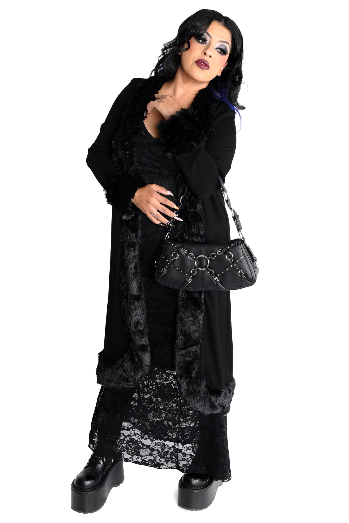 A woman modeling a gothic-style black costume consisting of a long faux-fur-trimmed cardigan over a black lace dress, paired with a black studded shoulder bag and black platform boots, against a plain white background.