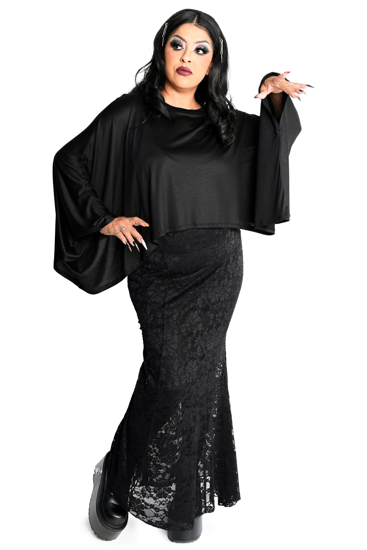 A woman modeling a black gothic-style outfit consisting of a satin cape top with wide bell sleeves and a black lace mermaid skirt, posed against a solid white background.