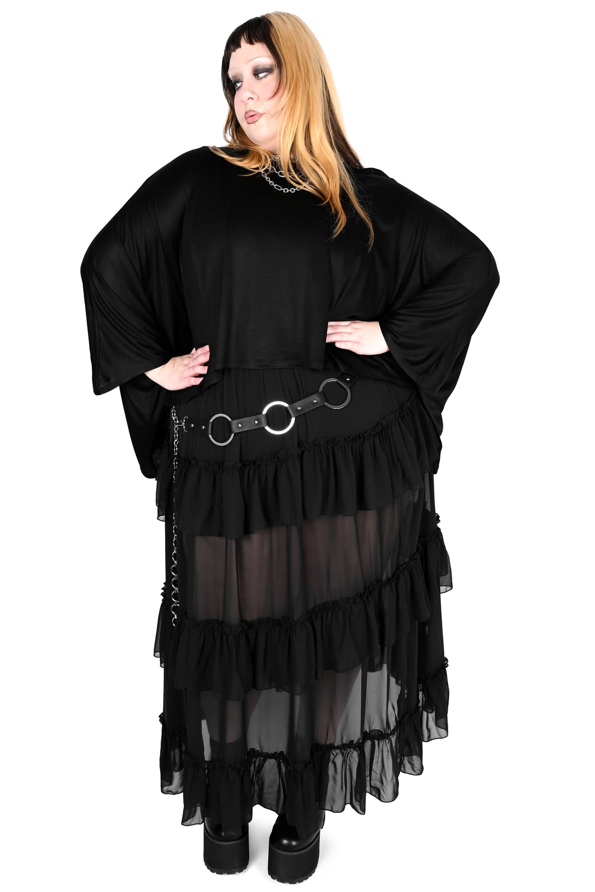 A woman modeling a black gothic-style outfit with wide sleeves, a sheer tiered skirt, and a black belt with silver rings, set against a plain white background.