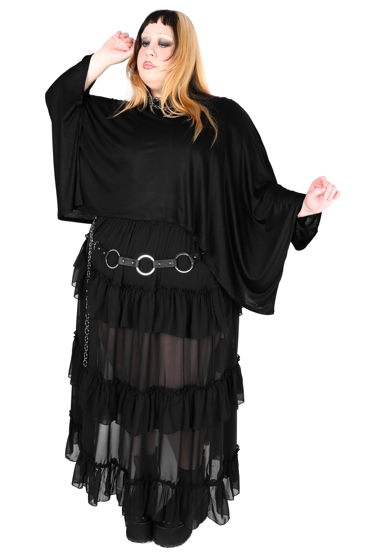 A woman modeling a black gothic-style top with wide draped sleeves, a tiered sheer skirt, and a black belt with large silver rings, against a plain white background.