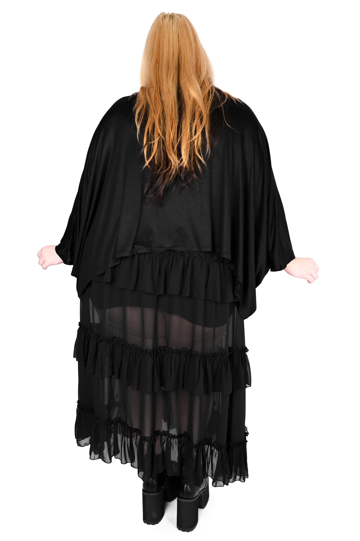 A woman viewed from behind wearing a top with wide draped sleeves over a ruffle skirt, standing against a plain white background.