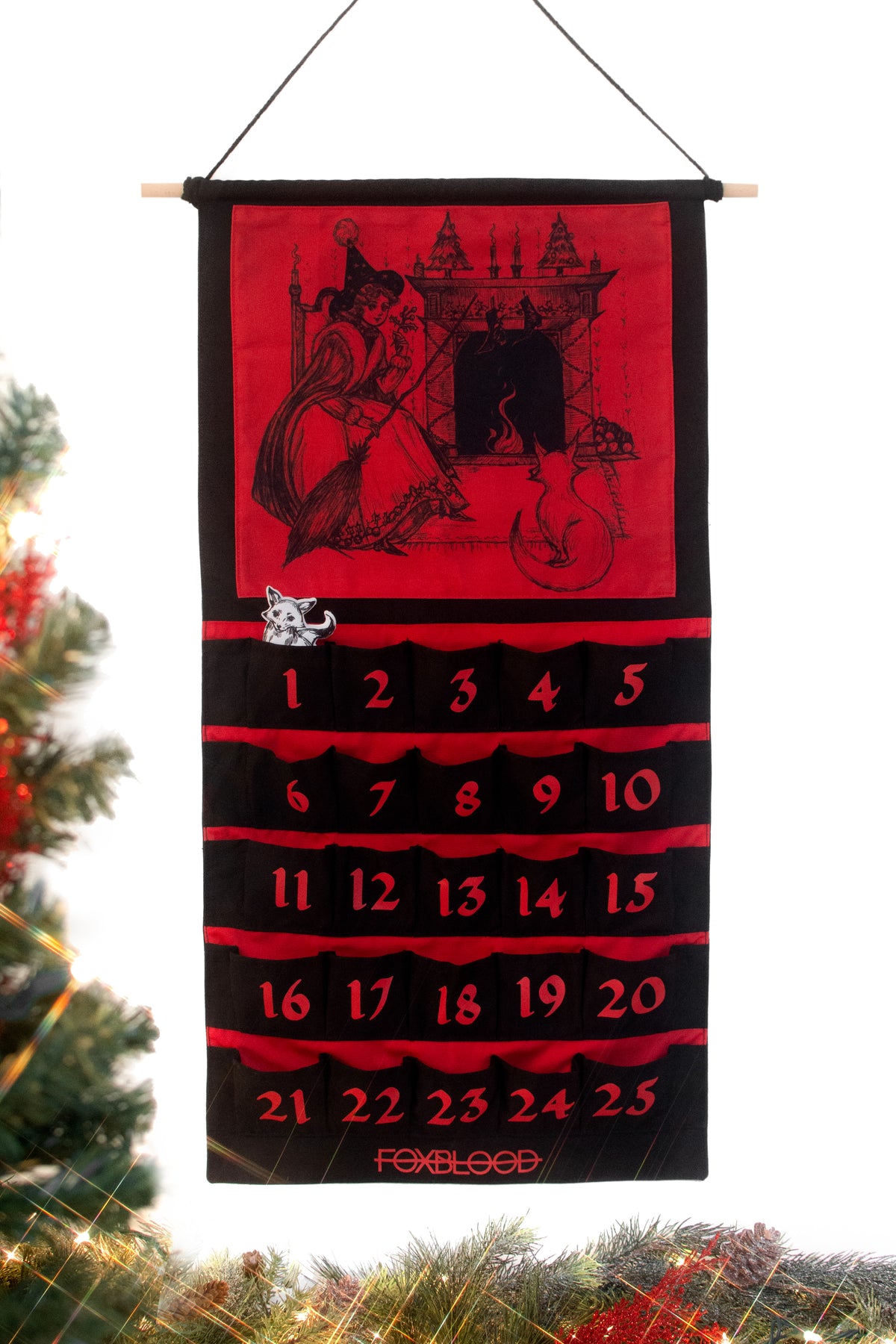 Large black and red canvas advent calendar hanging from a wooden dowel and black rope. Featuring 25 pockets for the 25 days leading up to christmas. The top half is a large drawing of a witch and her familiar, a fox, sitting by a holiday themed fire. FOXBLOOD logo in red at the bottom. A white fox market can be placed in the pockets to mark each day